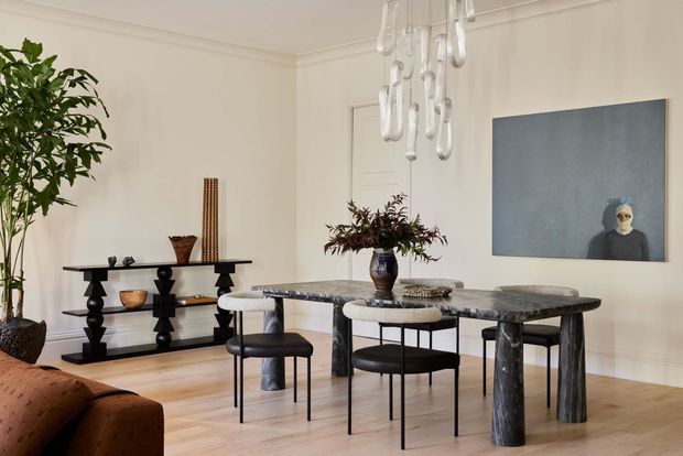 A refined dining room with pale timber flooring, warm off-white walls, and simple wall mouldings. A dark stone dining table with sculptural cylindrical legs is paired with black-framed chairs and stools. A black console with turned geometric supports sits against the wall, accompanied by a potted indoor tree and decorative objects. A large suspended glass pendant chandelier hangs above the table, while contemporary artwork adds a muted blue-grey accent.