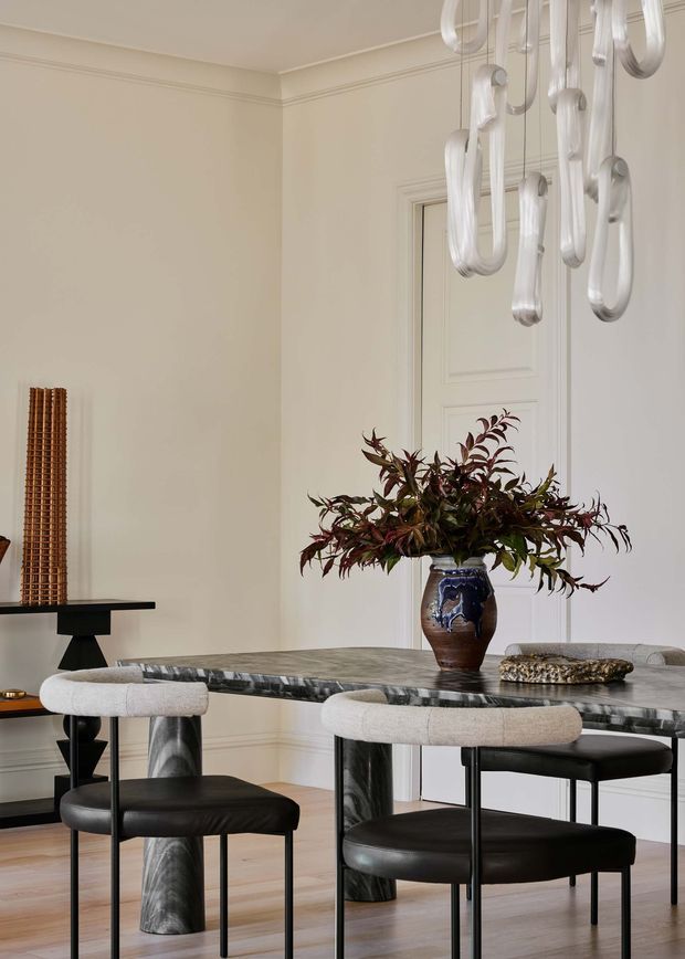 Elegant formal dining room with a sculptural marble dining table, upholstered dining chairs with black leather seats, a large floral arrangement, decorative wall panelling and trim, and a contemporary sculptural pendant light. The room has a restrained neutral palette with dark stone and black furniture accents, creating a refined, gallery-like atmosphere.
