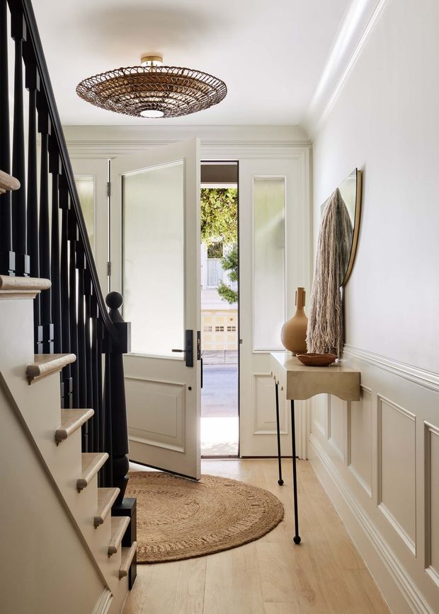 Bright, narrow residential entry hall with a staircase on the left, black vertical balustrade, pale timber flooring, and white decorative wall panelling and trims. A front door is open to the exterior, while a slim console table with a sculptural vase and textured wall art occupies the right wall. A woven pendant light and round jute rug add warmth to the refined space.