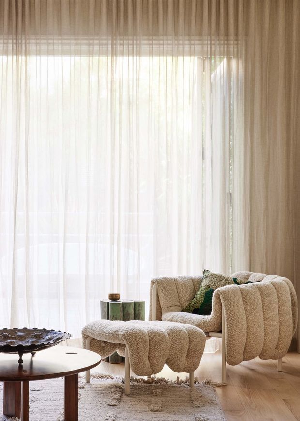 A softly lit contemporary living room with floor-to-ceiling sheer curtains, warm beige walls, pale timber flooring, and sculptural cream boucle seating. A matching armchair and ottoman sit beside a round wooden coffee table, with a textured rug and small green stone side table adding layered natural detail. The large glazed opening creates an airy, refined atmosphere.