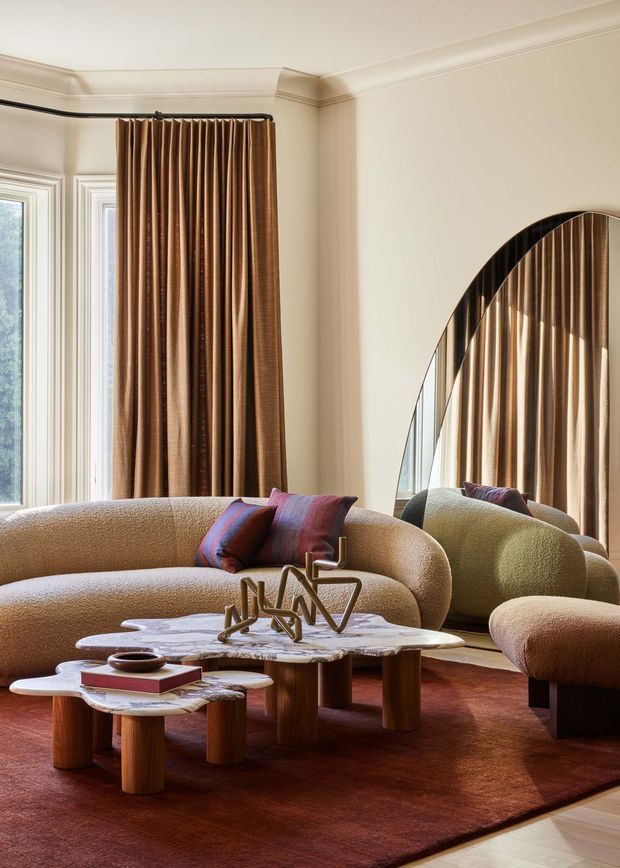 A refined living room with a curved textured sofa, coordinating rounded armchairs, layered organic-shaped marble coffee tables, and sculptural metal accessories. Floor-to-ceiling brown drapery frames tall windows, while a large arched mirror reflects the curtains and furniture. The room has cream walls with decorative ceiling and window mouldings, warm timber table bases, and a deep rust-red rug, creating a rich, sophisticated atmosphere.