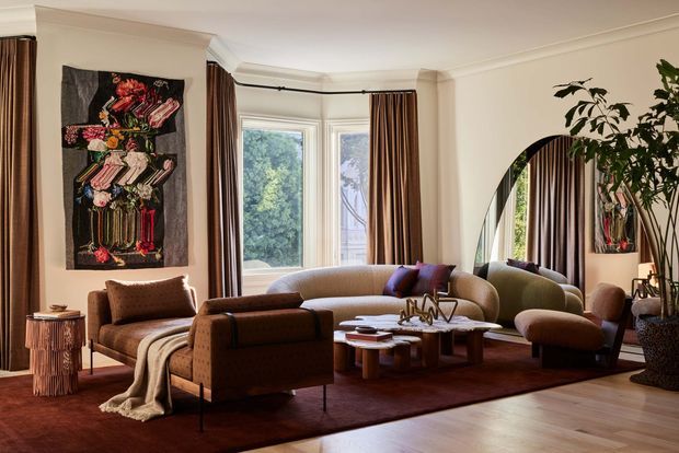 A refined living room with a warm, layered palette and period architectural detailing. The room features bay-style glazing with timber-toned curtains, decorative cornice moulding, a large arched mirror, a brown upholstered sofa and armchairs, a sculptural coffee table, dark area rug, timber flooring, an abstract floral wall hanging, and indoor planting.
