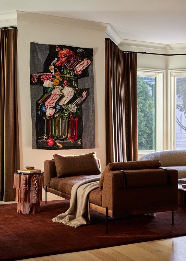 A warmly styled living room with two brown upholstered lounge chairs, a textured throw, a small sculptural side table, and a large burgundy area rug over timber flooring. Full-height dark brown curtains frame tall windows, while cream walls and elaborate traditional ceiling cornices surround a colourful floral textile artwork. The room combines classic architectural detailing with refined contemporary furniture and rich, moody colours.