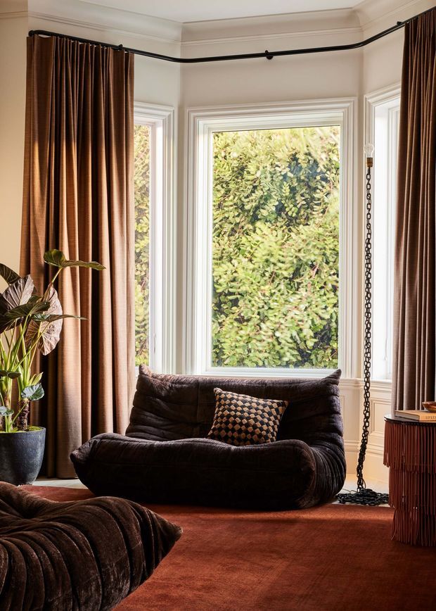 A richly styled sitting area in a bay-windowed room, featuring a deep dark brown lounge chair and ottoman, heavy brown floor-length curtains on a black ceiling-mounted track, a terracotta-brown carpet, patterned cushion, indoor plant, and a ribbed wooden side cabinet. White decorative wall panelling and moulded window trim frame the leafy garden view, creating a warm, moody, luxurious atmosphere.