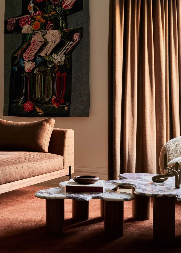 A warmly lit, richly layered living room vignette featuring a sculptural patterned sofa, a textured upholstered armchair, low irregularly shaped marble coffee tables with dark cylindrical legs, and a large floral textile artwork. Full-height pleated curtains frame the right side, while a rust-toned carpet and bronze, brown, cream, and muted pink tones create an intimate, curated atmosphere.