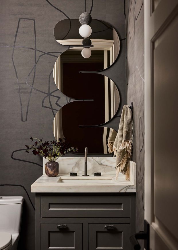 A dramatic, compact bathroom vanity viewed straight on. The room features a dark charcoal built-in vanity with raised-panel drawers, a pale veined stone benchtop and integrated basin, dark metal tapware, an organic sculptural mirror, and a striking globe pendant light. Charcoal patterned wallcovering, a draped textured towel, and a small vase add layered detail. A toilet is partially visible at the left edge and a doorway frames the right side.