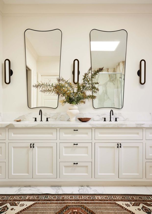 A refined bathroom vanity with a long double-sink cabinet, white raised-panel cabinetry, marble benchtop and splashback, black tapware, sculptural mirrors, and wall sconces. A patterned runner and restrained botanical arrangement add warmth to the otherwise light, classic space.