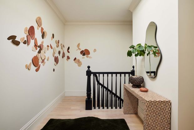 A refined residential stair landing with pale timber flooring, cream walls and detailed white cornices and skirting. A black-painted timber balustrade borders the stair opening, while a patterned console cabinet sits beneath an organically shaped wall mirror. Sculptural warm-toned wall art and a potted plant add decorative interest. The space is softly lit, calm and contemporary with traditional architectural detailing.