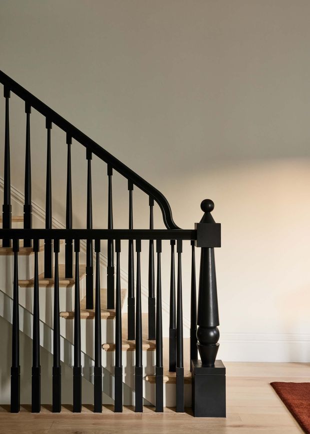 Interior view of a traditional stairway with a curved black painted balustrade, turned newel post, slender vertical spindles, and pale timber stair treads. The staircase rises along a light neutral wall with decorative trim, while pale timber flooring and part of a rust-coloured rug are visible at the base.