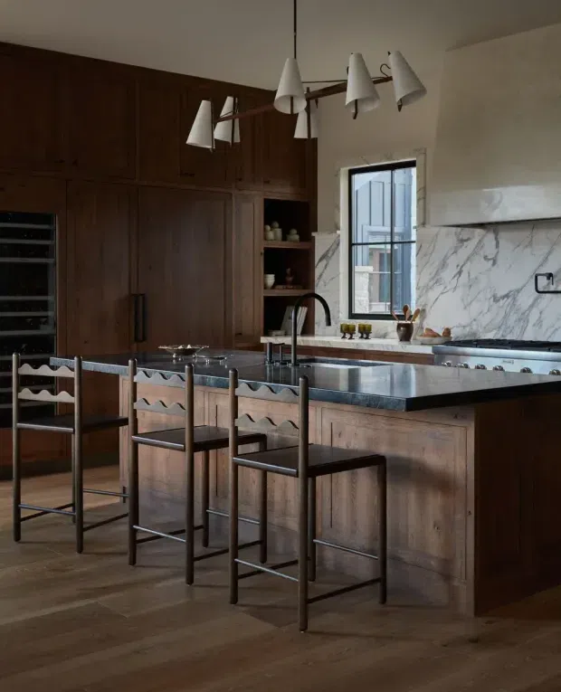 A warm, sophisticated kitchen with extensive dark timber cabinetry, a large central island, dark stone benchtops, and a dramatic white marble splashback with grey veining. A row of sculptural metal bar stools sits at the island, while a contemporary multi-light pendant hangs overhead. Natural light enters through the window, complementing the rich wood flooring and cabinetry.