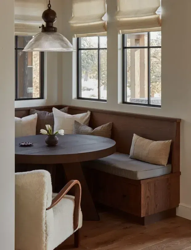A warm, intimate breakfast nook or dining corner with a built-in timber bench beneath a bank of black-framed windows. The space features a round dark wood table, upholstered cream armchair, neutral cushions, Roman blinds, timber flooring, and a large ribbed metal pendant light. The palette is earthy and refined, with natural wood and soft neutral textiles.