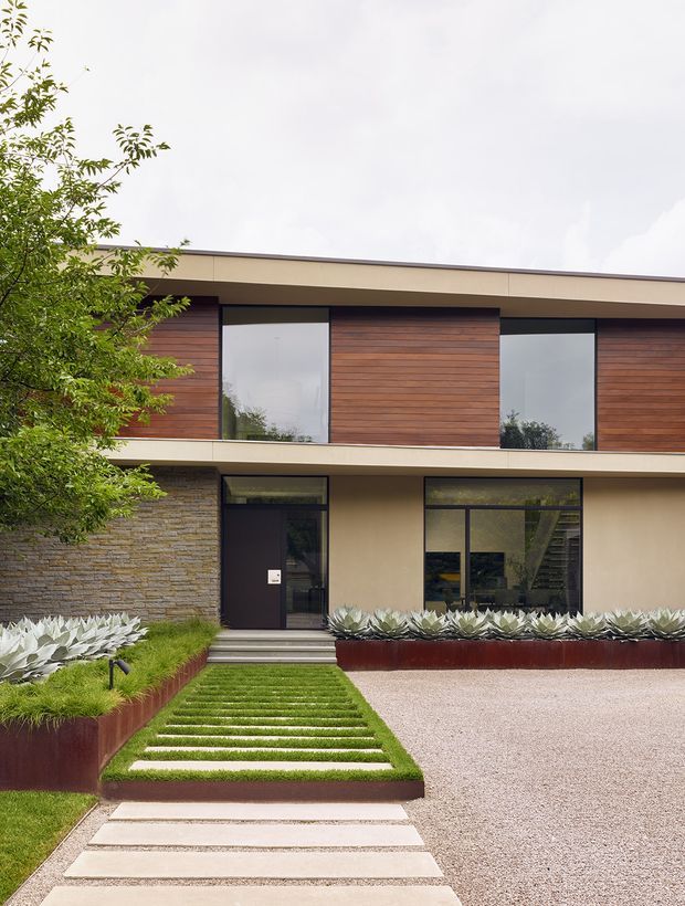 A contemporary two-storey residential facade with a broad low-pitched roof, clean horizontal lines, dark-framed glazing, warm horizontal timber cladding, and pale rendered wall surfaces. A textured stacked-stone wall anchors the lower level. The landscaped front approach includes a stepping-stone path through lawn, sculptural succulents, raised rust-toned planter edging, and a light gravel driveway.