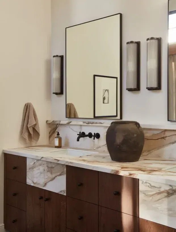 Elegant bathroom vanity with a long white and richly veined marble countertop and backsplash, dark timber drawer cabinetry, wall-mounted black tapware, framed mirror, fluted wall sconces, and a decorative ceramic vessel. Warm neutral walls and natural materials create a refined, classic atmosphere.