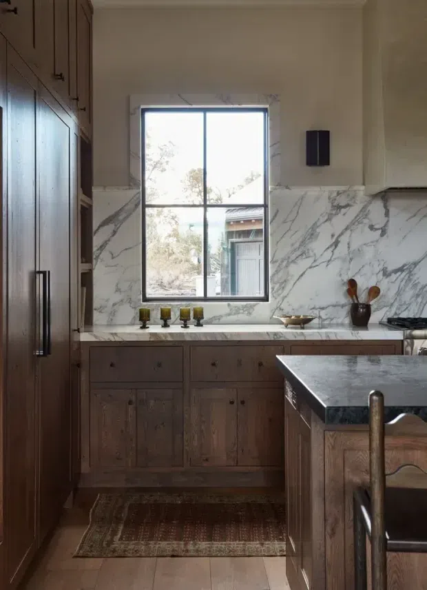A warm, traditional kitchen with dark stained timber cabinetry, a large white marble-look backsplash and benchtop, and a central island with a dark stone surface. A black-framed divided-light window provides natural light, while vintage-style hardware, a patterned runner, and wooden utensils add rustic character.
