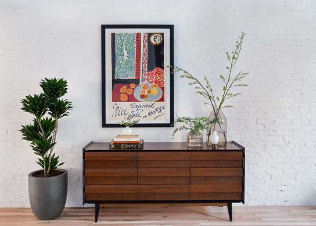 A styled interior vignette featuring a mid-century-inspired dark timber sideboard with tapered legs against a white-painted brick wall. A tall potted plant stands to the left, while a framed colorful artwork, books, and simple glass vases with leafy branches decorate the sideboard.