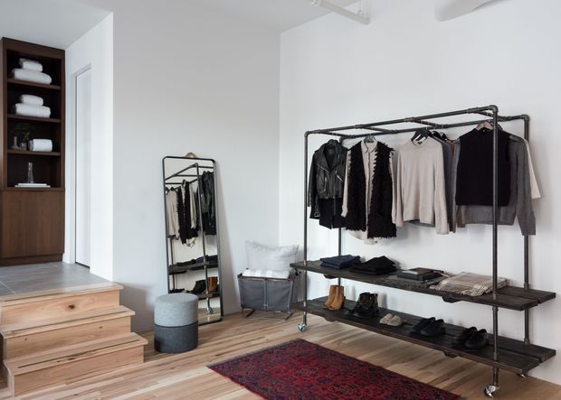 A bright, minimalist walk-in wardrobe or dressing area with exposed industrial-style black metal clothing racks, timber shelving, neatly arranged clothing and shoes, a full-length leaning mirror, wooden flooring, and timber stairs leading to a raised storage area. Built-in dark timber shelving with folded towels or linens is visible at the left.