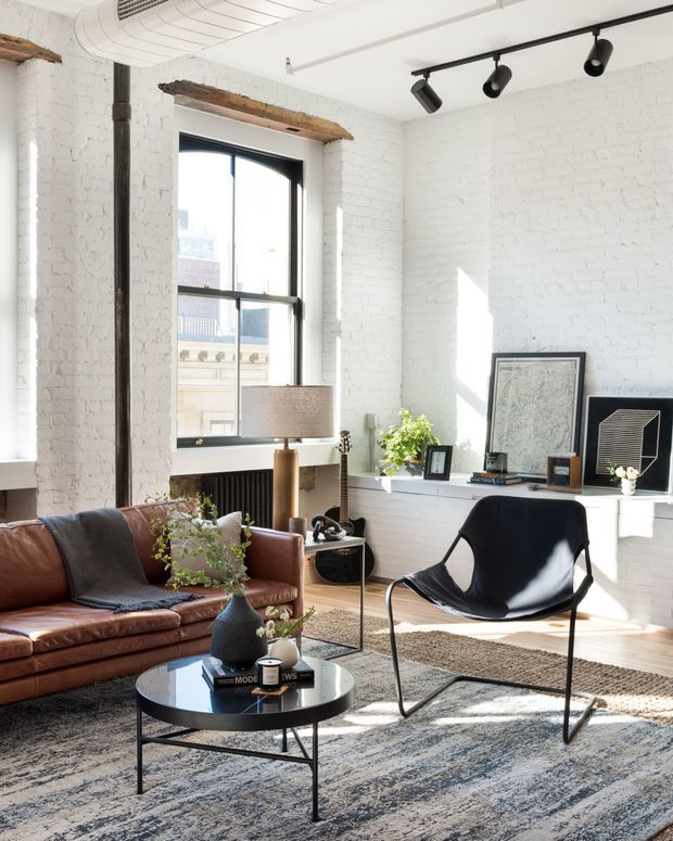 Bright loft-style living room with white-painted exposed brick walls, tall black-framed industrial windows, and a black track-lighting rail. The room is furnished with a cognac leather sofa, black sling chair, round black coffee table, floor lamp, patterned area rug, plants, and framed artwork. Exposed timber lintels and a visible radiator add character, while strong sunlight creates a warm, airy atmosphere.