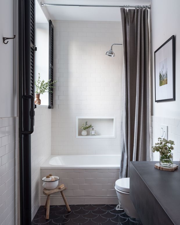 A bright, compact bathroom with white subway-tile walls, a built-in bathtub and shower, a dark patterned tile floor, and a partially screened toilet. A dark timber vanity with a white basin occupies the foreground, complemented by black louvred shutters, a dark shower curtain, framed artwork, and small plants. The room has a refined, modern-traditional feel with strong black-and-white contrast.