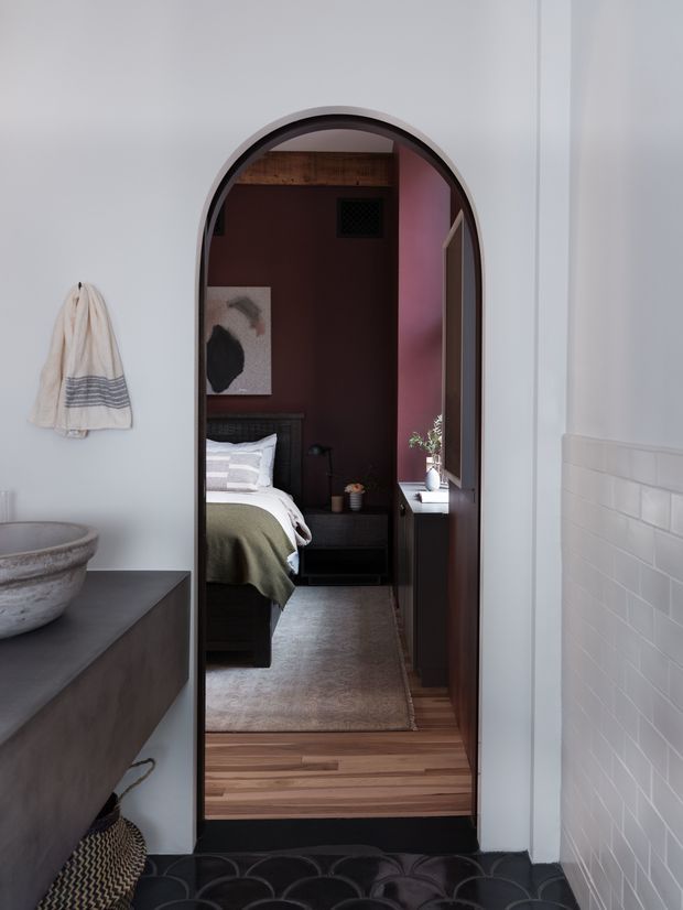 View through a tall arched doorway from a bright bathroom into a dark, richly coloured bedroom. The bathroom has white walls, white rectangular wall tiles, a dark vanity with a stone vessel basin, a hanging towel, and a patterned dark floor. The bedroom beyond features a deep burgundy accent wall, dark timber furniture and headboard, a partially visible bed with muted green and white bedding, timber flooring, and a textured rug. The image has a refined, moody, contemporary interior aesthetic with strong contrast between the two spaces.