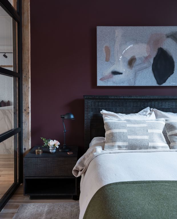 A moody, refined bedroom interior featuring a dark timber bed with a textured headboard, layered white and grey bedding, and a sage-green throw. A dark bedside table with a small lamp and flowers sits against a deep burgundy wall. Black-framed glazing with a rustic timber surround is visible on the left, and abstract artwork hangs above the bed.