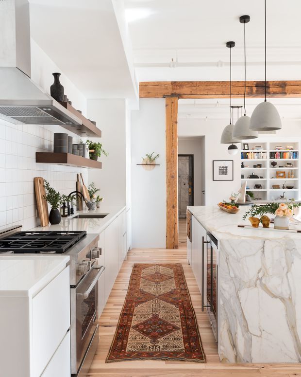 Bright, open-plan kitchen with white cabinetry, a marble waterfall island, white tiled backsplash, timber flooring, exposed rustic timber beams and posts, open shelving, and multiple pendant lights. A dining or living area with shelving is visible beyond the kitchen, while a patterned runner adds warmth to the central walkway.