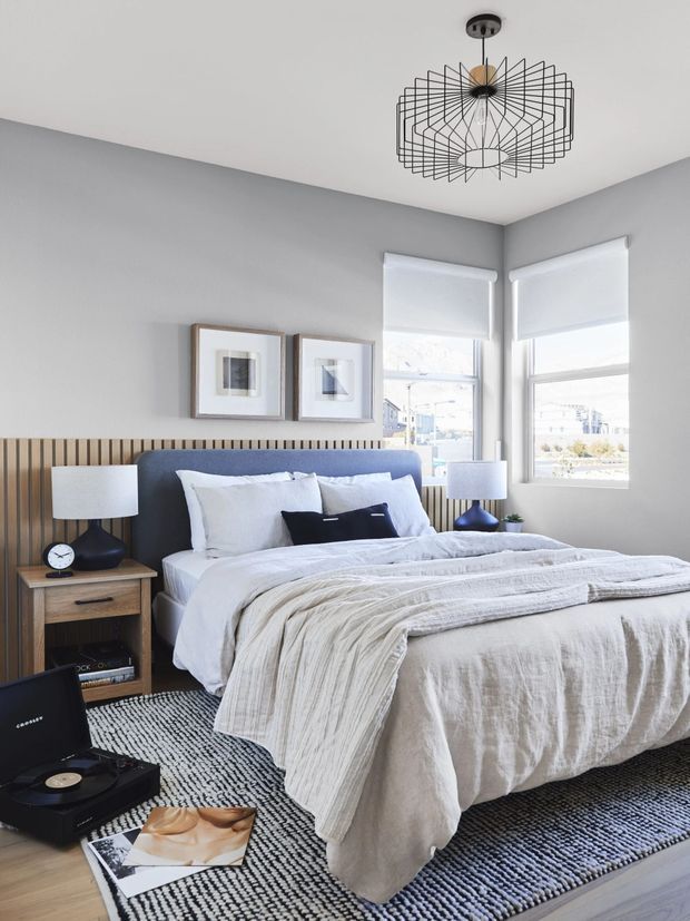 A bright, contemporary bedroom with a low upholstered blue-grey bedhead, layered white and pale grey bedding, and a timber slatted feature wall behind the bed. Two large corner windows have white roller blinds, bringing in abundant natural light. Matching bedside tables with dark sculptural lamps flank the bed, while framed artwork, a black-and-white textured rug, a record player, and magazines add a refined but relaxed character. A geometric black wire pendant light provides a striking ceiling feature.