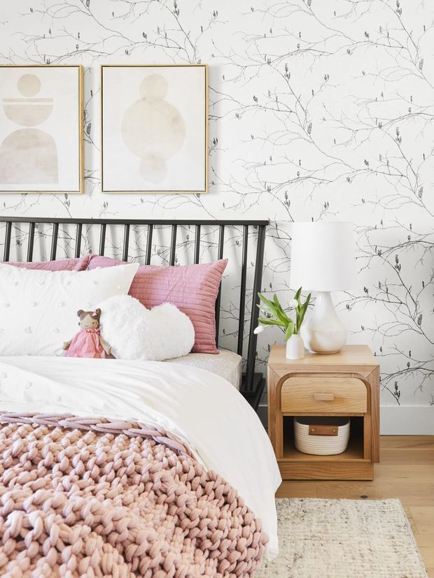A bright, softly styled bedroom featuring a black metal bed frame, white bedding, blush-pink cushions, a chunky blush knit throw, and a small plush toy. The room has delicate botanical wallpaper, pale timber flooring, framed neutral abstract artwork, and a compact timber bedside table with a white lamp and small plant.