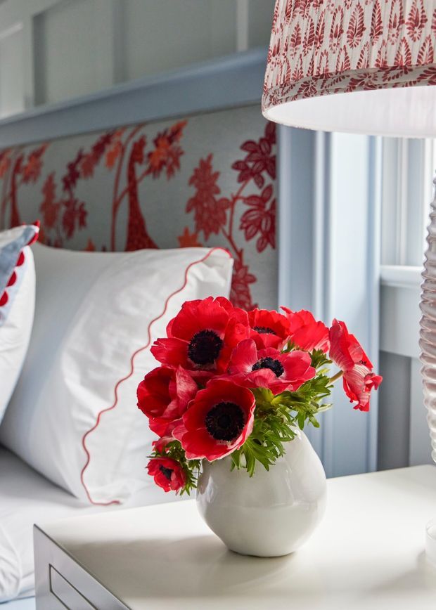 Close-up interior view of a refined bedroom corner featuring a white bedside table, a rounded white vase with vivid red flowers, white bedding with red piping, and a pale blue wall upholstered or covered with a bold red botanical pattern. A patterned fabric lampshade and layered window trim add decorative detail.