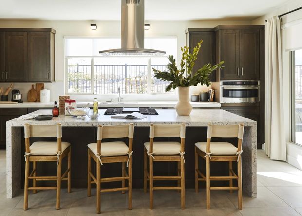 Bright, spacious contemporary kitchen centered on a large stone island with four timber-framed upholstered bar stools. Dark wood cabinetry, integrated stainless-steel appliances, a broad window with outdoor views, white curtains, and a large floral arrangement create a warm, polished family kitchen setting.