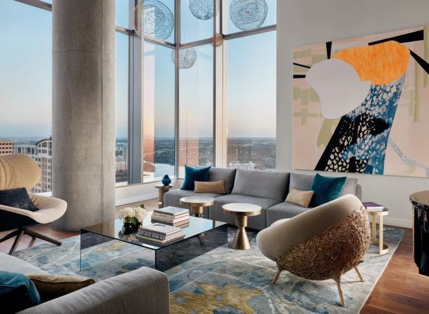 A sophisticated high-rise living room with floor-to-ceiling glazing and expansive city views. The room features a large grey sectional sofa, sculptural lounge chairs, round metallic coffee tables, a glass coffee table, a patterned area rug, colourful cushions, and a large abstract artwork. A prominent exposed concrete column and visible window framing contribute to the contemporary architectural character, while hanging spherical decorative lights add visual interest.