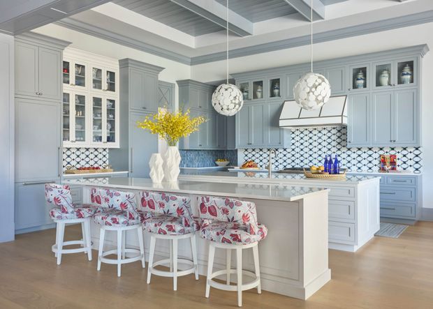 Bright, spacious kitchen with a large central island, pale grey shaker-style cabinetry, glass-front display cupboards, and a white patterned tile splashback. The island includes multiple floral upholstered stools, while two spherical pendant lights hang above. Light timber flooring, marble-look benchtops, and a coffered ceiling create a refined, traditional coastal-inspired aesthetic.