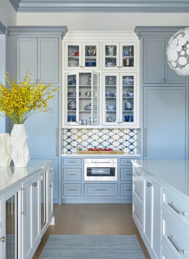 A refined blue-grey kitchen with a compact central cooking station, glass-front display cabinetry filled with blue-and-white ceramics, pale stone benchtops, and a decorative patterned tile splashback. Symmetrical tall cabinetry frames the room, while a white sculptural vase with yellow branches and a spherical pendant add visual accents. The space has detailed traditional-style mouldings and a calm, light palette.