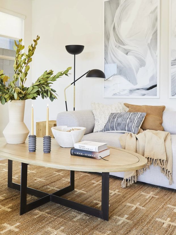 Bright, softly styled living room featuring a light neutral sectional sofa, pale timber oval coffee table with a black metal base, layered cushions and throw, woven rug, sculptural black floor lamp, large abstract artwork, and a tall ceramic planter with leafy branches. The room has a warm, relaxed contemporary feel with abundant natural light.