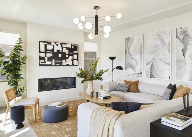 Bright, refined contemporary living room with a large cream sectional sofa arranged around a round timber coffee table, a built-in linear fireplace, abstract wall art, and a sculptural globe pendant light. The room features pale textured wall panelling, light neutral furnishings, a woven rug, black accent furniture and lighting, and abundant natural light.