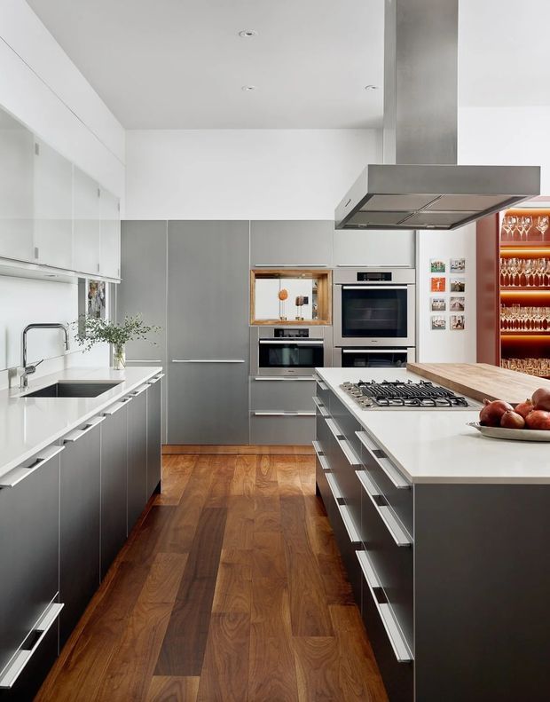 A sleek contemporary galley kitchen with a central island, white and grey flat-panel cabinetry, integrated stainless-steel appliances, a white stone benchtop, and a large stainless-steel rangehood. Warm timber flooring contrasts with the cool cabinetry. A built-in open wine-glass display is visible at the rear, with a sink and tap along the left wall and a gas cooktop on the island.