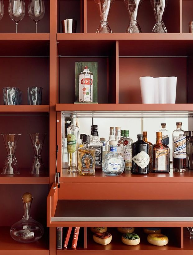 Close-up view of a built-in home bar with rich reddish-brown cabinetry and open display shelving. The shelves hold wine glasses, tumblers, decanters, cocktail glasses, spirits, and bar accessories, with a mirrored or reflective back panel illuminating the bottle display. Lower shelving includes books and decorative objects.