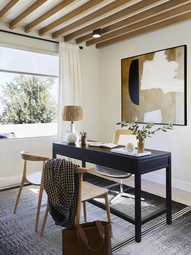 Bright, refined home office with a black rectangular desk, mixed wood and upholstered chairs, neutral textiles, and minimalist decorative accessories. A large abstract artwork hangs on the white wall, while broad glazing provides abundant daylight. The ceiling features exposed warm timber beams with white infill panels, creating a distinctive architectural detail.