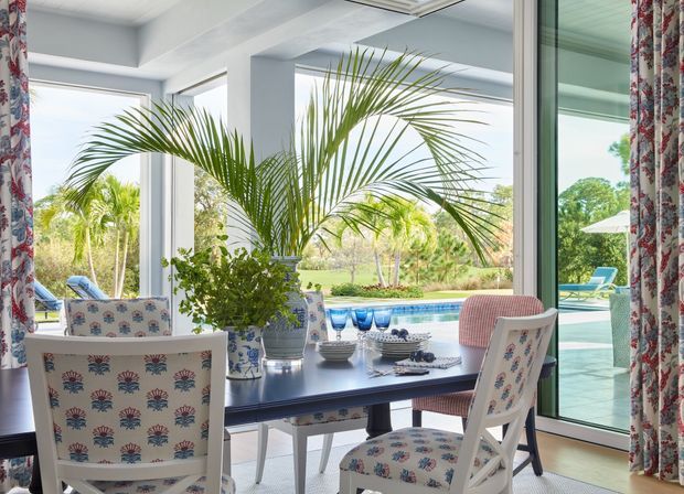 Bright, airy dining area with a dark rectangular dining table and patterned upholstered chairs, positioned beside expansive floor-to-ceiling glazing. Tropical palms, a swimming pool, and landscaped grounds are visible outside. Floral patterned curtains frame the windows, while white structural columns and a pale ceiling create a light coastal feel.