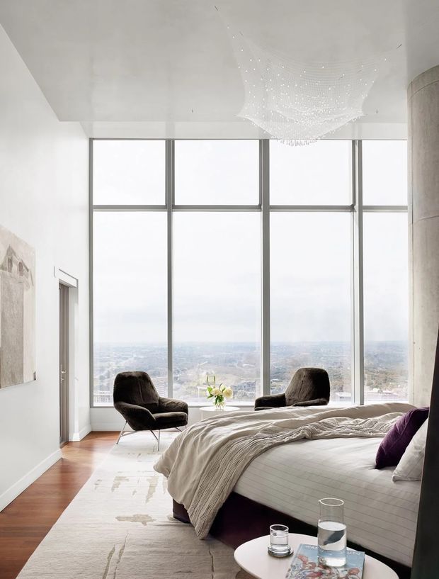 Bright, spacious high-rise bedroom with floor-to-ceiling grid-framed windows overlooking a distant cityscape. The room features a large upholstered bed with layered neutral bedding, dark sculptural lounge chairs, a small round bedside table, timber flooring, a pale area rug, white walls and ceiling, and a delicate suspended sculptural light fitting. A curved concrete column frames the right side of the room.