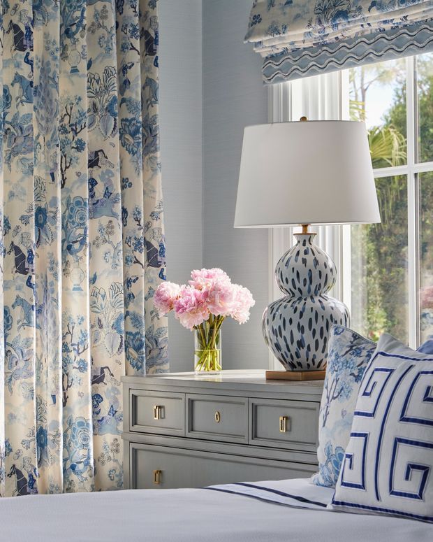 A refined bedroom vignette featuring blue-and-white patterned curtains, a matching Roman blind, a grey painted bedside chest with brass hardware, and a sculptural blue-and-white ceramic lamp. A bed with blue geometric bedding and a vase of pink flowers complete the layered, classic interior.