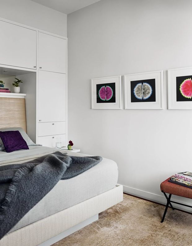 A compact, neatly styled bedroom with a light upholstered bed, grey throw, purple accent pillow, built-in white cabinetry and open shelving, framed botanical artwork, and a small upholstered bench. The room has pale grey walls, soft neutral furnishings, and a warm beige carpet, creating a calm contemporary interior.