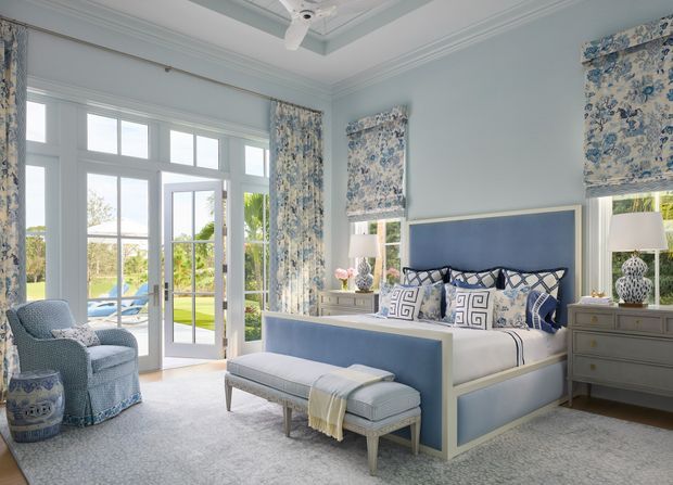Bright, elegant bedroom with a pale blue upholstered bed, matching bench and armchair, patterned blue-and-white Roman blinds and bedding, light carpet, and traditional decorative wall and ceiling mouldings. Large glazed French doors open toward a pool and landscaped garden, while matching bedside lamps and a grey dresser complete the refined coastal-inspired scheme.