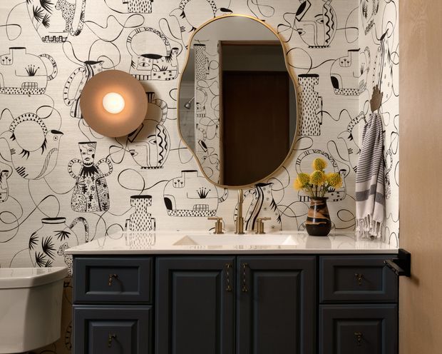 A compact, highly styled bathroom vanity with a dark blue-black traditional paneled cabinet, white basin, brass tapware and hardware, and an oval brass-framed mirror. The walls feature playful black-and-white illustrated wallpaper, complemented by a sculptural globe wall light and a small floral arrangement.
