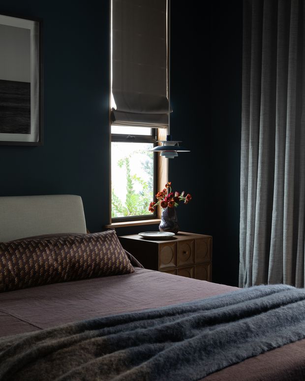 A moody, richly layered bedroom interior with deep charcoal-blue walls, a neatly made bed, patterned accent pillow, soft grey throw, sculptural bedside cabinet, floral arrangement, framed artwork, and a narrow window dressed with a Roman blind and full-length curtains. The room has an intimate, refined atmosphere with strong contrast between dark walls and filtered daylight.
