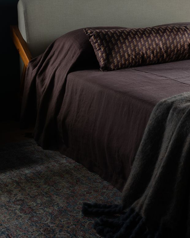 Close-up, moody bedroom scene featuring a neatly made bed with dark plum-brown linen bedding, a patterned lumbar cushion, a grey upholstered headboard, and a dark throw draped over the foot. A muted patterned rug is visible in the foreground, with warm timber detail at the bedside.