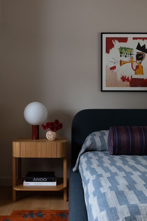 A warmly styled bedroom vignette featuring a dark upholstered bed with blue patterned bedding and a striped bolster cushion. Beside it is a compact mid-century-inspired timber bedside table with open shelving, a spherical white table lamp, and a small vase of red flowers. A framed abstract artwork hangs on the taupe-grey wall, with timber flooring and a partial rust-toned rug visible.
