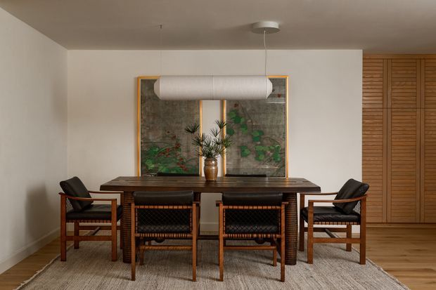 A warm, refined dining room with a rectangular dark timber dining table surrounded by six woven-back timber armchairs. A linear pendant light hangs above the table, with botanical artwork or screens behind it and full-height timber louvred cabinetry or wall panelling to the right. Neutral plaster walls, timber flooring, and a natural woven rug create a cohesive mid-century-inspired interior.