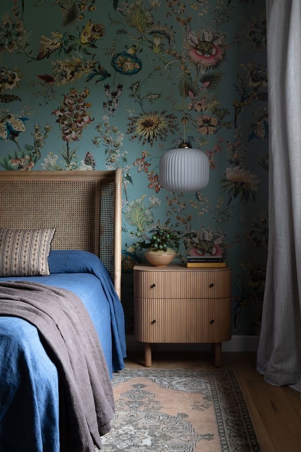 A richly styled bedroom featuring a tall cane-panelled bedhead, deep blue bedding, patterned neutral throw and cushion, and a small fluted wood bedside cabinet. The walls are covered in dramatic dark teal botanical wallpaper, complemented by a ribbed globe pendant light, sheer grey curtains, a patterned rug, and warm timber flooring. The room has an intimate, eclectic and nature-inspired atmosphere.