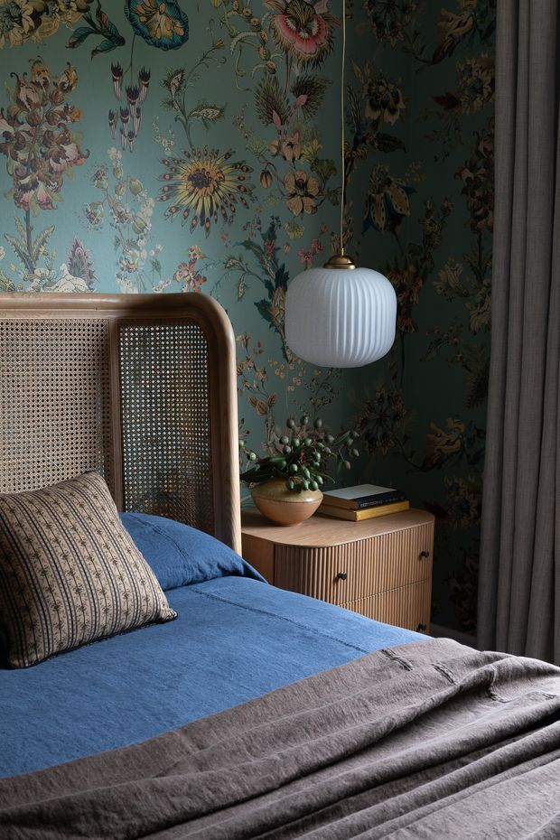 A moody, richly layered bedroom featuring dark teal floral wallpaper, a woven cane bedhead with rounded timber framing, blue linen bedding, a grey throw, and a fluted timber bedside cabinet. A ribbed white pendant light, potted plant, books, and full-height dark curtains complete the refined, botanical interior.