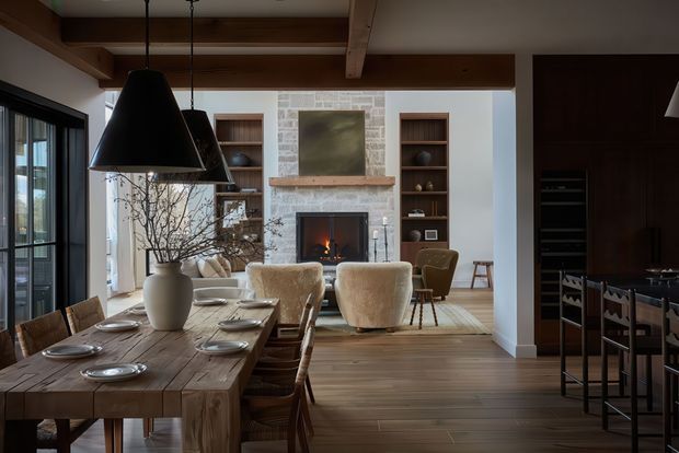 A refined open-plan dining and living area with a substantial rustic timber dining table set for guests, sculptural pendant lights, exposed timber ceiling beams, and wide dark-framed glazing. Beyond the dining area is a living room with a stone fireplace, built-in timber shelving, upholstered lounge chairs, and a warm fire. Dark timber cabinetry and bar seating are visible to the right.